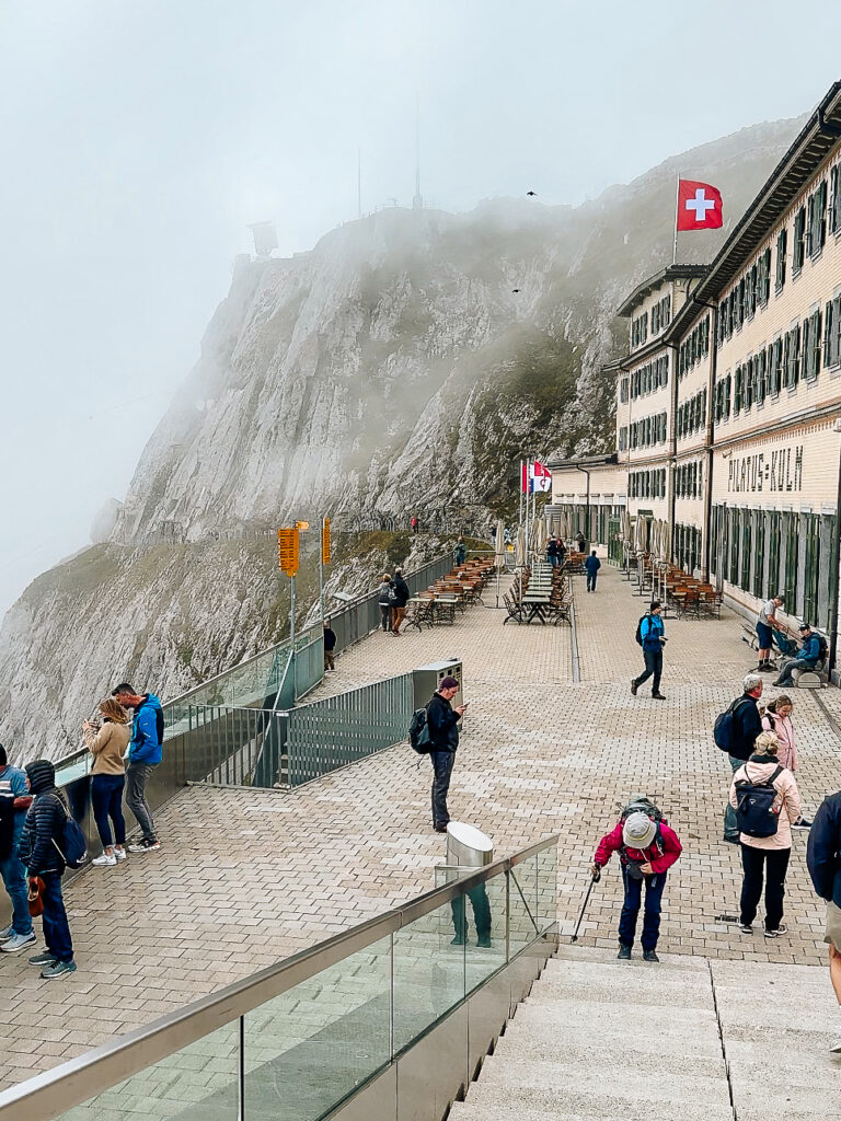 Tourists walking along the terrace of Pilatus Kulm with mountain cliffs disappearing into the fog.